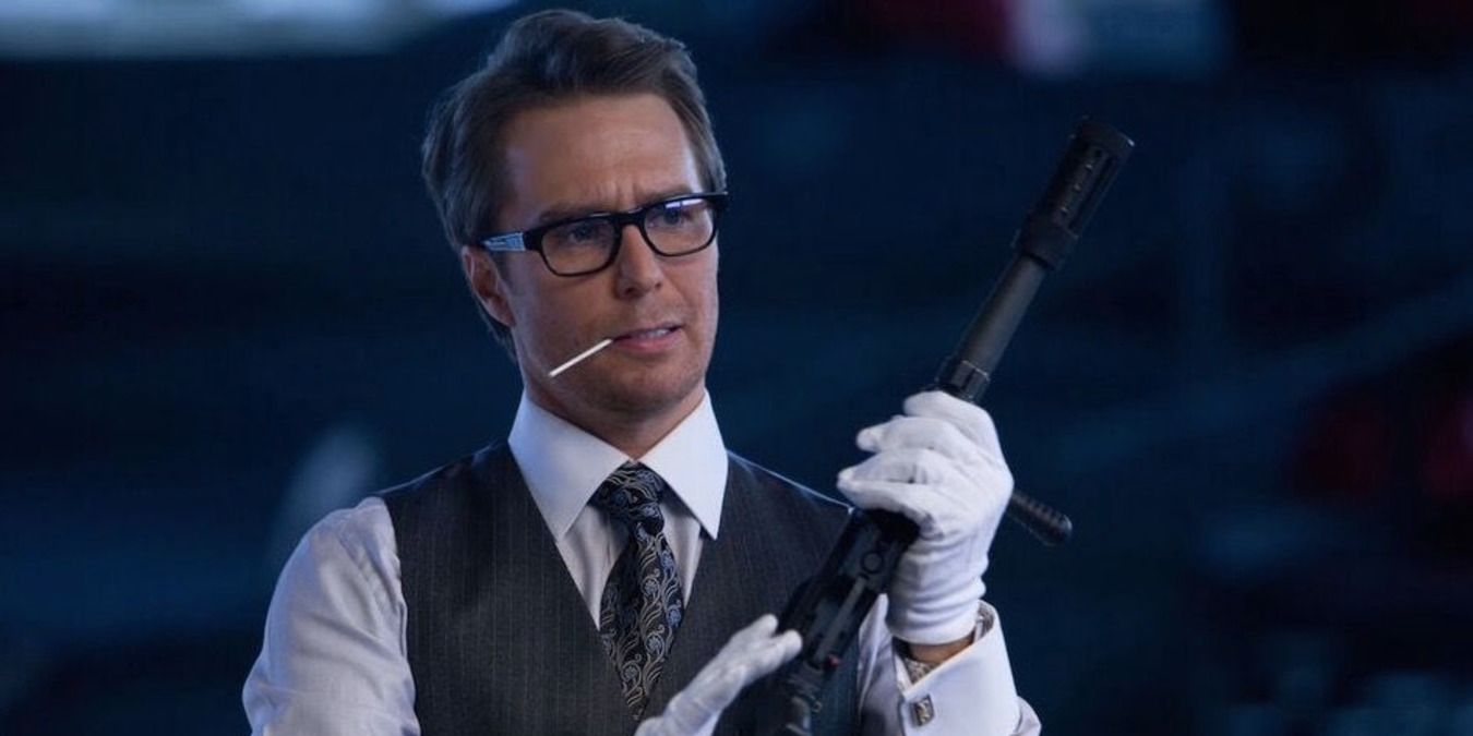 Justin Hammer holding a gun in the hanger wearing gloves and a suit vest