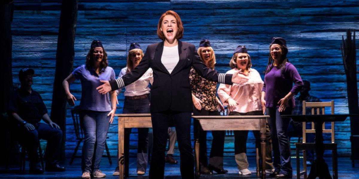 a woman in a suit singing with air hostesses around her in Come From Away