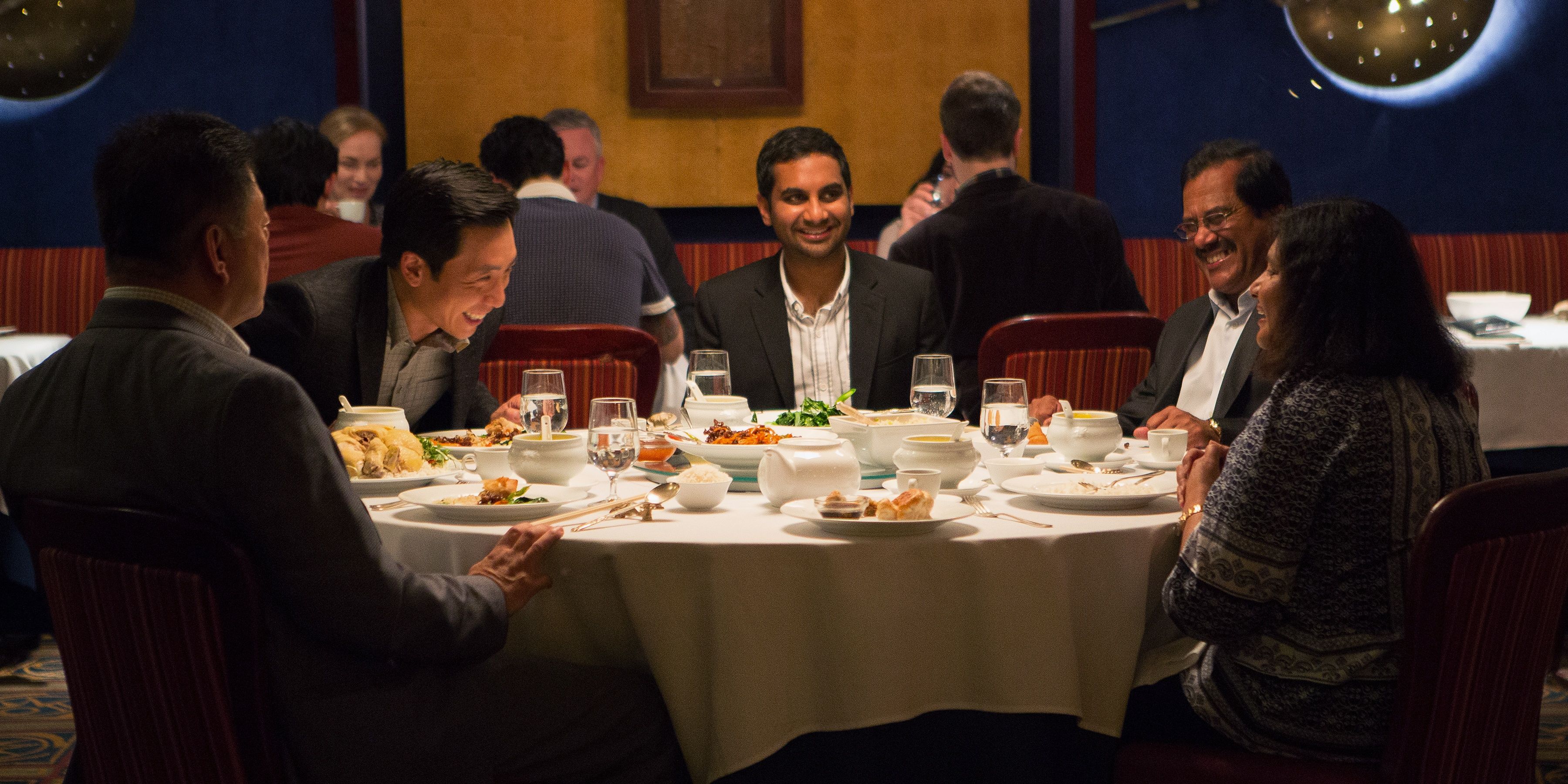 Dev sitting on a table and laughing with his friend and parents in Master of None Cropped
